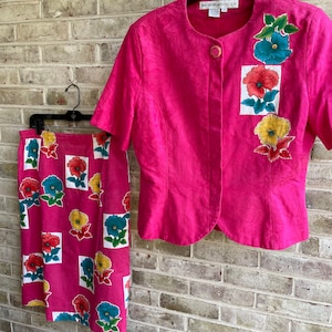 May include: A pink skirt and jacket set with a floral print. The skirt has a straight silhouette and the jacket has a button closure. The fabric has a textured surface.