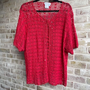 May include: A bright red crochet cardigan featuring a delicate openwork pattern, short sleeves, and a single button closure at the neckline. The garment includes a white brand label at the inner collar and is displayed on a black hanger.