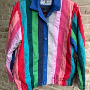 May include: A striped jacket with a blue collar and a button closure. The jacket is made of different colored fabrics, including pink, red, green, blue, and white. The label on the jacket reads "Marguerite Rebel".
