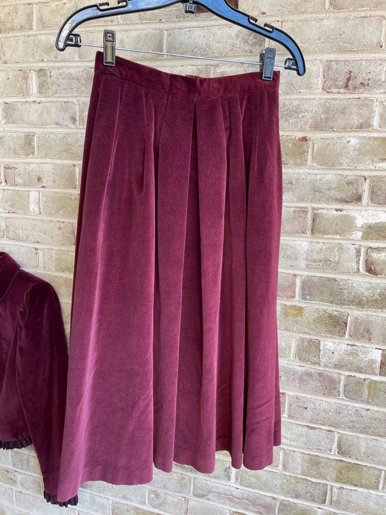 May include: A burgundy velvet skirt with pleats. The skirt is hanging on a hanger and has a simple design.