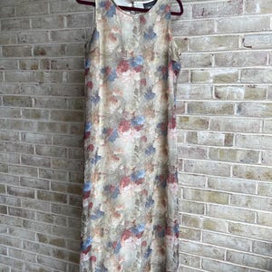 May include: Sleeveless floral print dress in beige, red, and blue hues. The dress is hanging on a burgundy hanger against a brick wall. The dress features a round neckline and a flowing, floor-length design, perfect for a summer day.