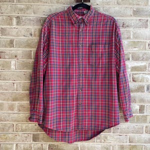 May include: A long-sleeve button-down shirt featuring a red, navy blue, and yellow plaid pattern. The shirt includes a pointed collar, a single chest pocket on the left side, and a curved hemline. The front is secured with a vertical row of buttons.
