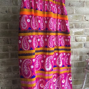 May include: A long, pink skirt with a paisley pattern in white, orange, and blue. The skirt has a wide waistband and a flowing silhouette.