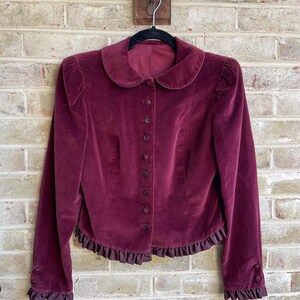 May include: A burgundy velvet jacket with a Peter Pan collar, long sleeves with ruffled cuffs, and a ruffled hem.