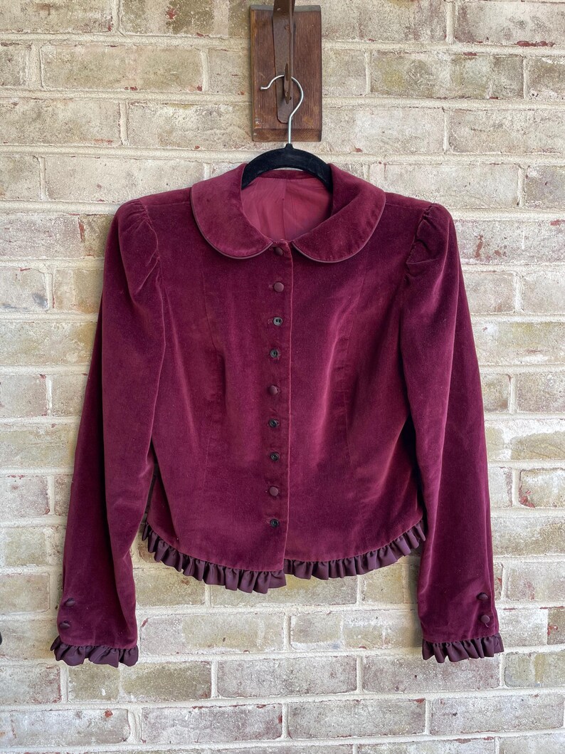 May include: A burgundy velvet jacket with a Peter Pan collar, long sleeves with ruffled cuffs, and a ruffled hem.