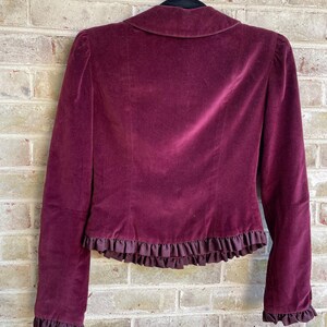May include: A burgundy velvet jacket with a ruffled trim around the bottom and sleeves. The jacket is hanging on a wooden hanger.