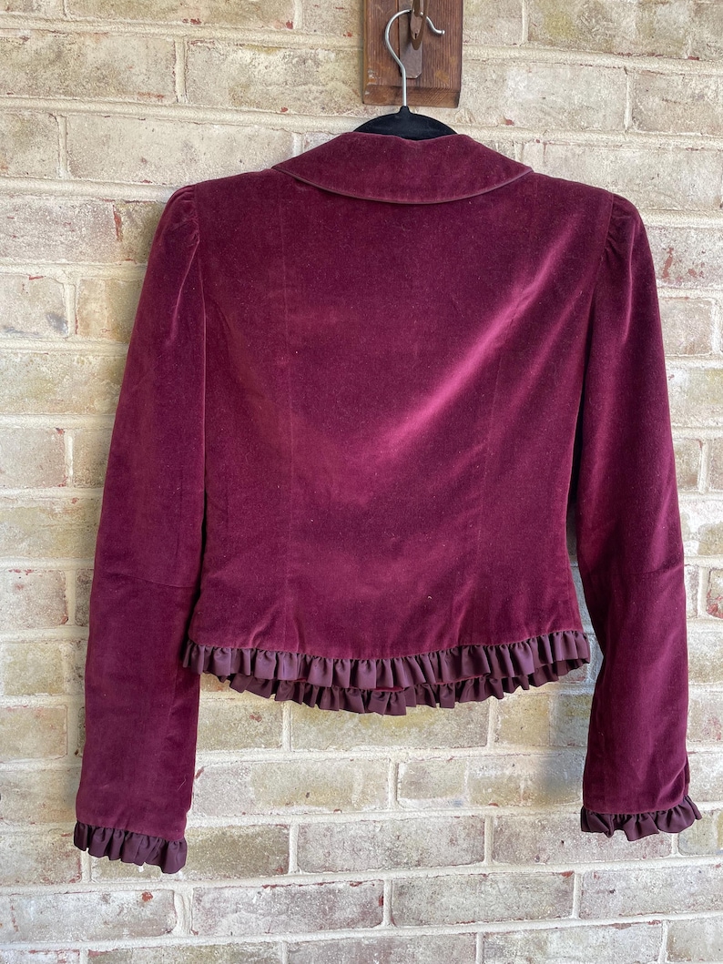 May include: A burgundy velvet jacket with a ruffled trim around the bottom and sleeves. The jacket is hanging on a wooden hanger.