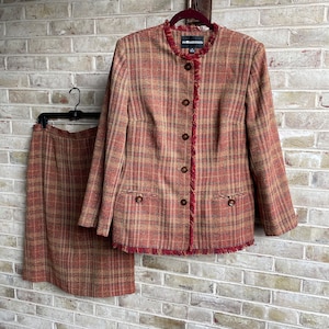 Puede incluir: Un traje de falda de cuadros rojos, marrones y beige con ribete de flecos. La chaqueta tiene cuello mandarín y cierre de botones. La falda es de largo hasta la rodilla.