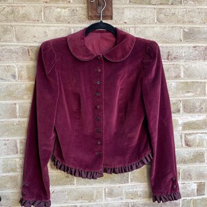 May include: A burgundy velvet jacket with a peter pan collar, long sleeves, and a ruffled hem. The jacket has a button-up front closure.