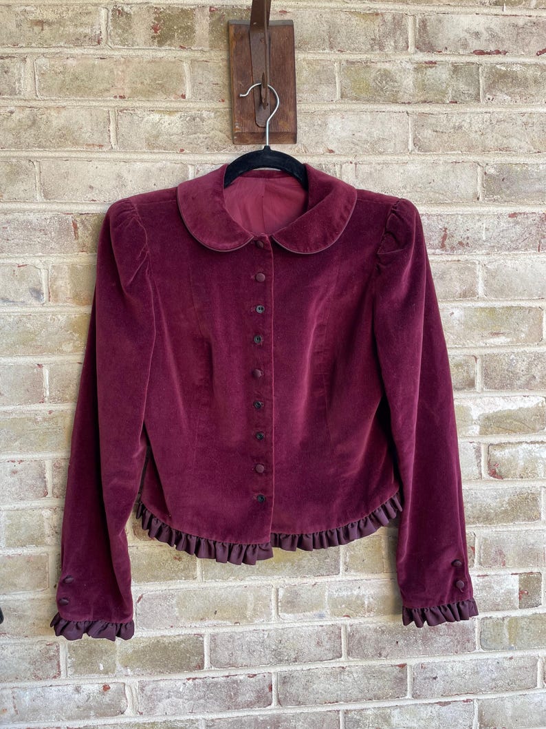May include: A burgundy velvet jacket with a peter pan collar, long sleeves, and a ruffled hem. The jacket has a button-up front closure.