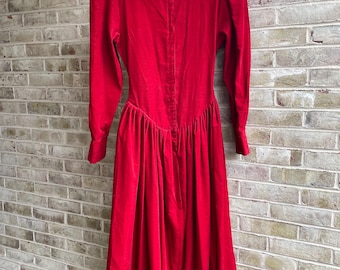 May include: A vibrant red dress with a high neckline and long sleeves hangs against a brick wall. The dress features a fitted bodice, a gathered waist, and a flowing skirt. The fabric appears to be a soft velvet.