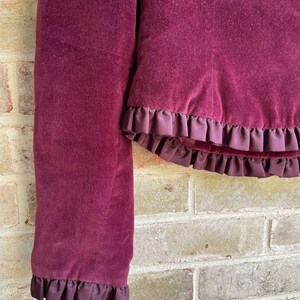 May include: A burgundy velvet jacket with a ruffled trim. The jacket has long sleeves and a cropped length.