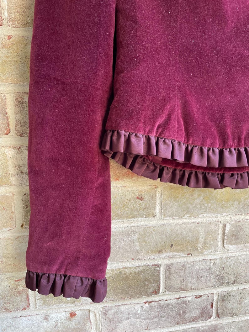 May include: A burgundy velvet jacket with a ruffled trim. The jacket has long sleeves and a cropped length.