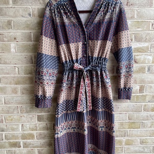 May include: A long-sleeved, blue and beige patterned dress with a button-up front and a tie belt. The dress has a V-neck and a gathered waist.