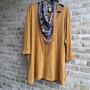 May include: A mustard yellow V-neck top with three-quarter length sleeves, paired with a triangular scarf featuring a black, white, and gold patterned print and long fringe trim, all displayed on a black hanger.