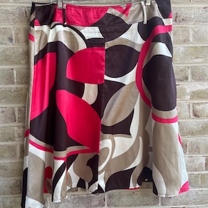 May include: A-line skirt with a vibrant abstract pattern. The skirt features a mix of colors, including brown, pink, beige, and white, in a flowing, geometric design. The skirt is hanging on a hanger against a brick wall.