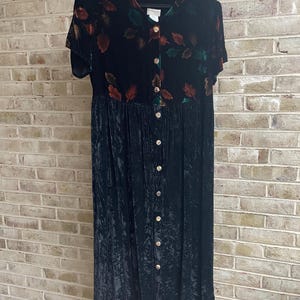 May include: A black velvet dress with short sleeves and a button-down front. The bodice features a pattern of colorful leaves in shades of red, green, and brown. The dress is hanging on a black hanger against a brick wall.