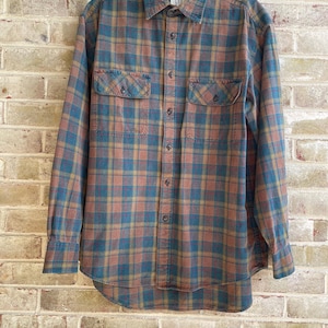 May include: A long-sleeve button-down shirt featuring a plaid pattern in muted shades of teal, rust orange, and mustard yellow. The shirt includes a standard collar, a front button placket, and two chest pockets with buttoned flaps.