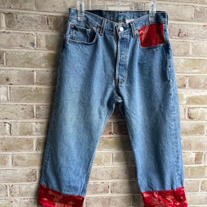 May include: Blue denim jeans with red and gold embroidered trim on the cuffs and back pocket.