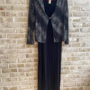 May include: A black two-piece dress set. The set includes a long, sleeveless dress and a matching long-sleeved jacket. The jacket is black with silver-colored embellishments. The dress and jacket are hanging on a brown hanger against a brick wall.