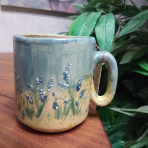 May include: Handmade ceramic mug with a blue and yellow floral design. The mug features a cylindrical shape with a handle and is decorated with blue and white flower accents. The mug is approximately 4 inches tall and is suitable for hot or cold beverages.