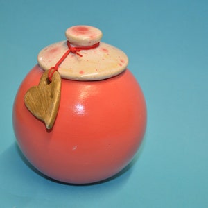 Può includere: Un piccolo vaso rotondo in ceramica di colore corallo con un coperchio bianco e un'etichetta di legno a forma di cuore legata con un nastro rosso.