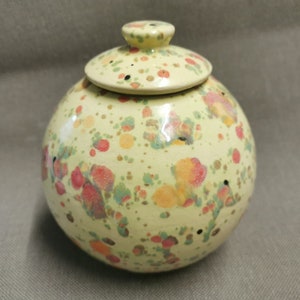 Può includere: Un piccolo vaso rotondo in ceramica con un coperchio. Il vaso è decorato con un motivo maculato di punti rossi, arancioni, gialli, verdi e marroni su uno sfondo giallo chiaro.