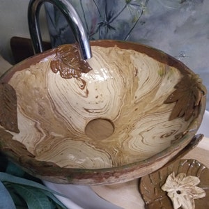 May include: A ceramic sink with a brown and white marbled design. The sink has a leaf-shaped design around the rim and a small drain hole in the center. The sink is sitting on a wooden surface.