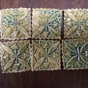 Könnte beinhalten: Sechs quadratische Keramikfliesen mit erhabenem geometrischem Design. Die Fliesen weisen ein zentrales Sternmuster mit blattartigen Formen und Dreiecken in Grün- und Beigetönen auf. Die Kanten haben eine gewellte Verzierung.