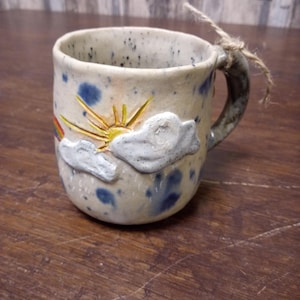 Puede incluir: Una taza de cerámica hecha a mano con un diseño caprichoso. La taza presenta un sol, nubes y un arcoíris, con motas azules sobre un fondo color crema. El asa es de un tono gris más oscuro y una cuerda está atada alrededor del asa.