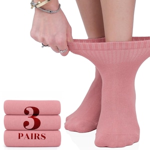 May include: A set of three pairs of solid dusty rose crew socks. The socks feature a ribbed cuff at the top and a smooth knit texture throughout. A stack of the folded socks is displayed on the left with the text 3 PAIRS printed in dark red.