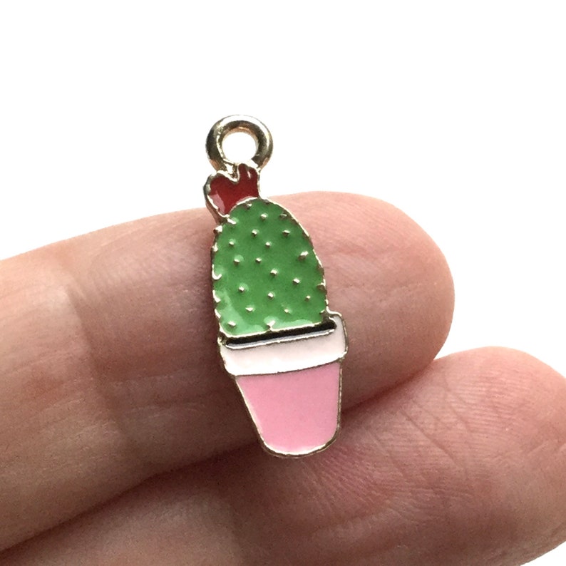 Pode incluir: Encanto de metal dourado com um cacto verde com uma flor vermelha no topo, plantado em um vaso rosa.