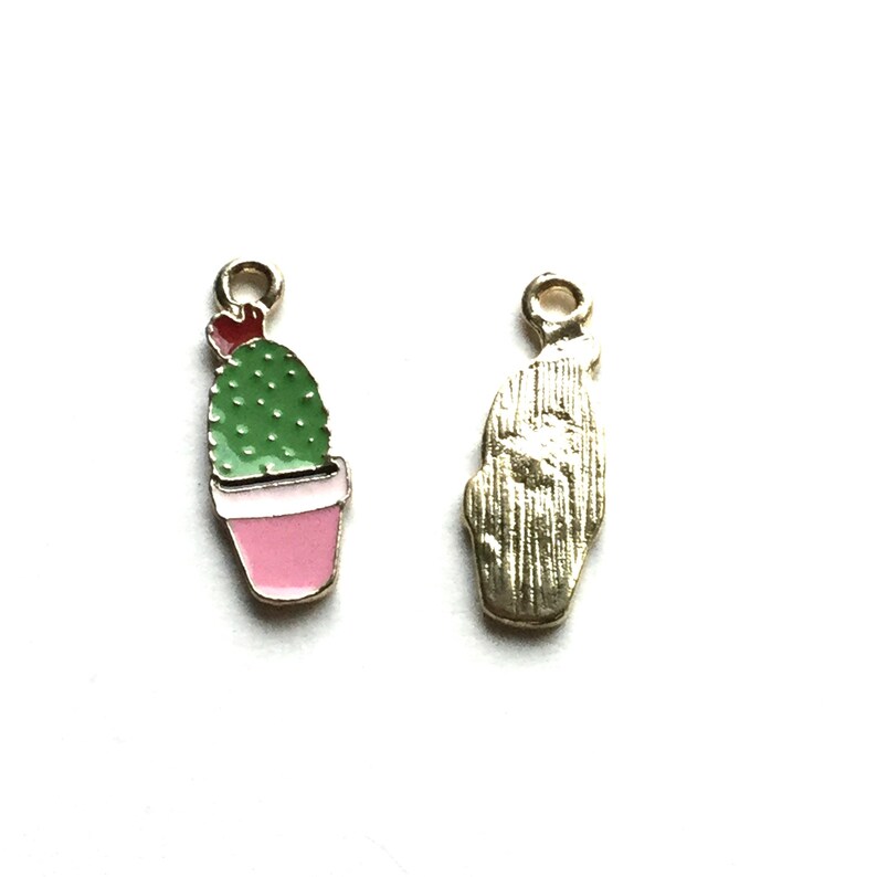 Pode incluir: Dois pingentes de metal, um com um cacto verde em um vaso rosa e o outro com um cacto dourado texturizado. Ambos os pingentes t&ecirc;m um la&ccedil;o para prender a um colar ou pulseira.