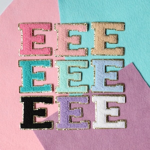 May include: Nine felt letter patches in various colors, each with a gold outline. The colors include pink, light blue, turquoise, lavender, white, and black. The letters are all the letter "E".