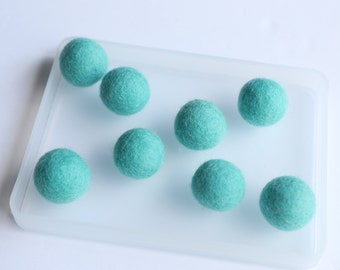 May include: Eight teal felt balls arranged on a clear plastic tray. The balls are all the same size and are made of a soft, fuzzy material.