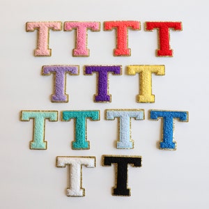 May include: A set of 12 iron-on felt letter patches in various colors, including pink, red, yellow, purple, blue, and white. Each patch features a gold outline and the letter "T" in a fuzzy, textured style.