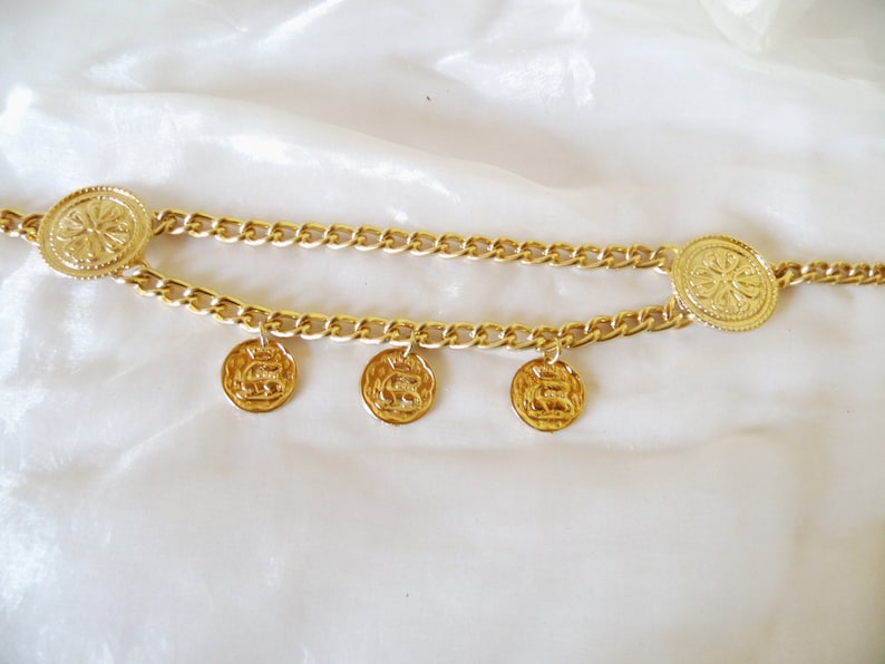 May include: A gold chain belt with three round, decorative charms. The chain is linked together with a thick, gold chain.