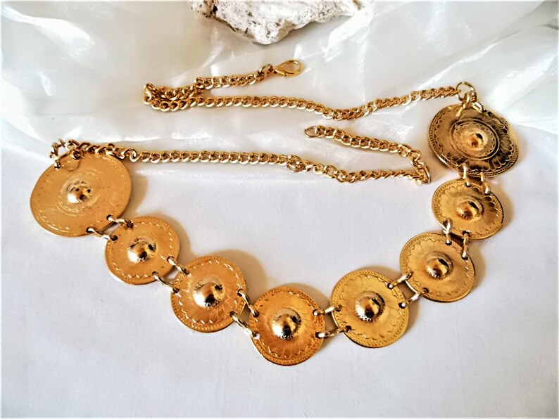 Puede incluir: Un cintur&oacute;n de cadena dorada con m&uacute;ltiples medallones redondos decorativos de color dorado. El cintur&oacute;n est&aacute; hecho de una cadena de eslabones y tiene un cierre de broche.