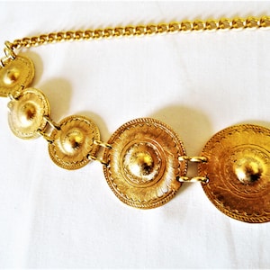 Puede incluir: Un cintur&oacute;n de cadena dorado con cinco medallones redondos de metal adornados. Los medallones tienen un dise&ntilde;o texturizado en relieve.
