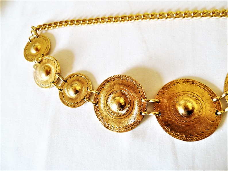 Puede incluir: Un cintur&oacute;n de cadena dorado con cinco medallones redondos de metal adornados. Los medallones tienen un dise&ntilde;o texturizado en relieve.