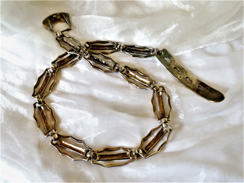 May include: A silver chain belt with a decorative clasp and a long, rectangular pendant. The chain is made of interlocking links that are shaped like elongated ovals.