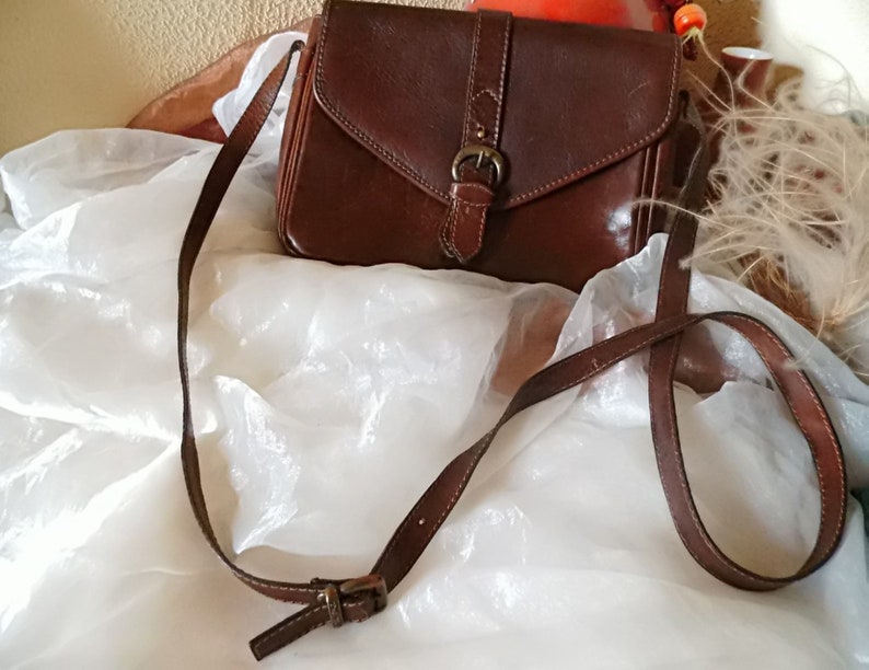 May include: A brown leather crossbody bag with a flap closure and a metal buckle. The bag has a long adjustable strap.
