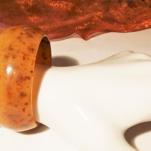 Op de afbeelding: Een brede, oranje-bruine armband met donkerbruine spikkels. De armband wordt gepresenteerd op een witte mannequinarm, waarbij het gladde, gepolijste oppervlak wordt benadrukt. Het sieraad heeft een klassiek, afgerond ontwerp.