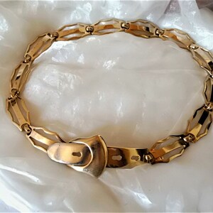 May include: A gold-toned metal belt with a decorative buckle. The belt is made of linked, interlocking pieces that form a chain-like design.