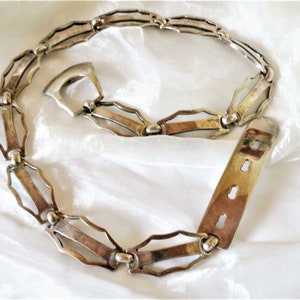 May include: A silver chain belt with a geometric design. The belt has a buckle with a keyhole design and a sliding closure.