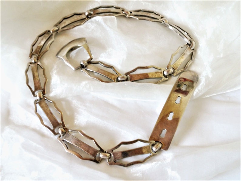 May include: A silver chain belt with a geometric design. The belt has a buckle with a keyhole design and a sliding closure.