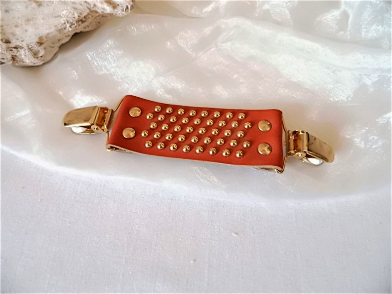 May include: A brown leather strap with gold studs and two gold metal clips. The strap is designed to hold items together.