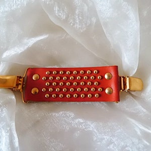 May include: A red leather hair clip with gold metal accents and a studded design. The clip has a rectangular shape and is decorated with small gold studs.