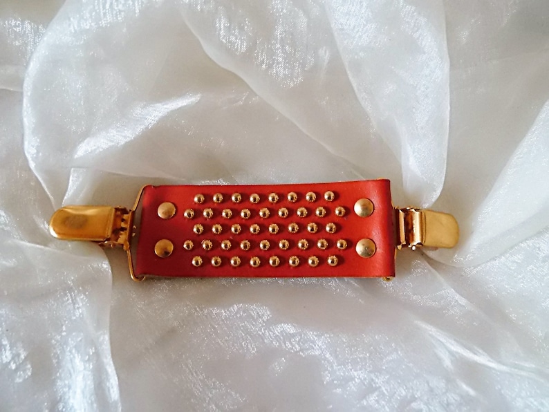 May include: A red leather hair clip with gold metal accents and a studded design. The clip has a rectangular shape and is decorated with small gold studs.