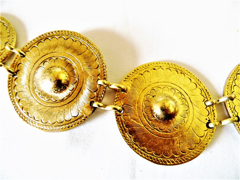 Puede incluir: Un cintur&oacute;n dorado con medallones redondos, ornamentados y decorativos. Cada medall&oacute;n tiene un centro elevado y un dise&ntilde;o de c&iacute;rculo conc&eacute;ntrico.